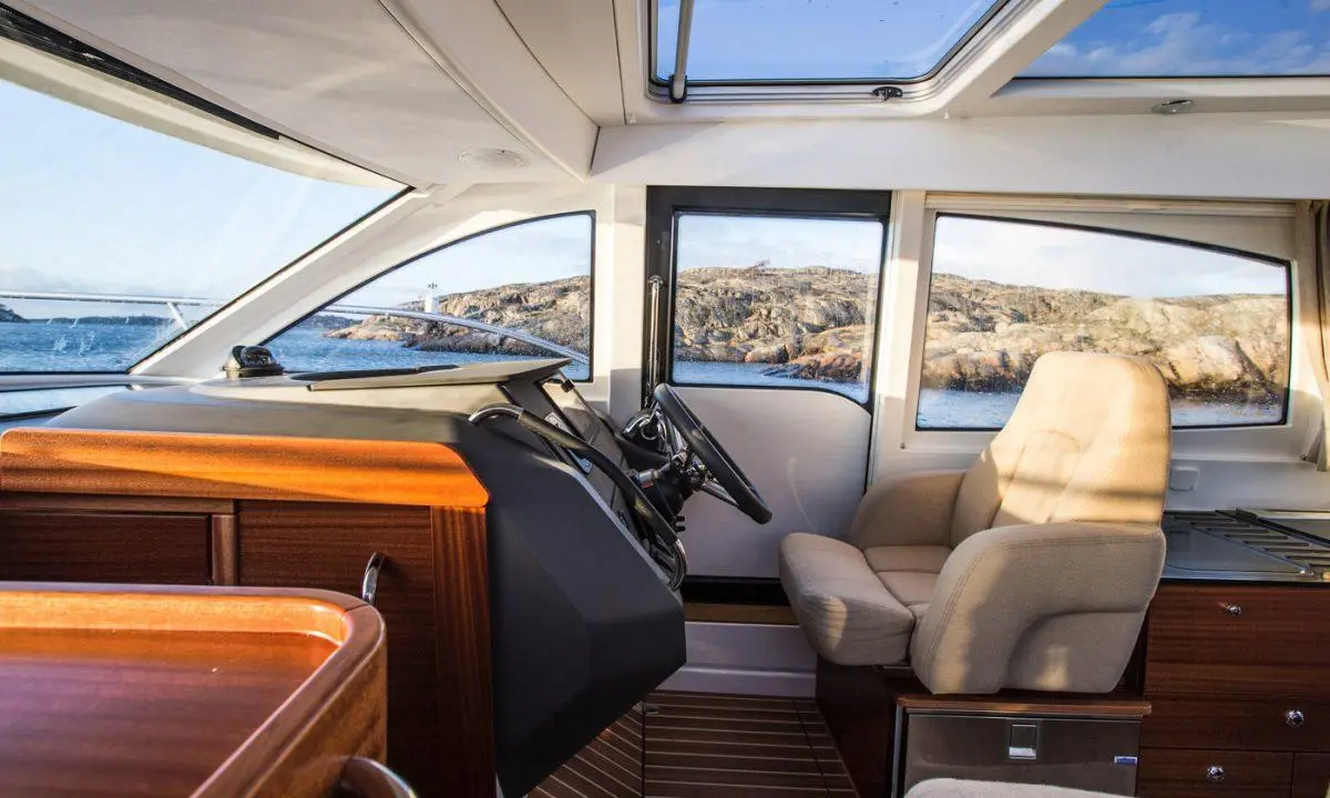 Interior of a boat cabin with a steering wheel and comfortable seating, featuring scenic views of the water and rocky landscape.